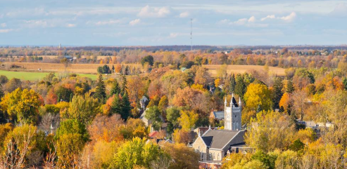 Aerial view of a quaint town amidst vibrant autumn foliage. A church with a tall steeple stands out, surrounded by fields and trees.