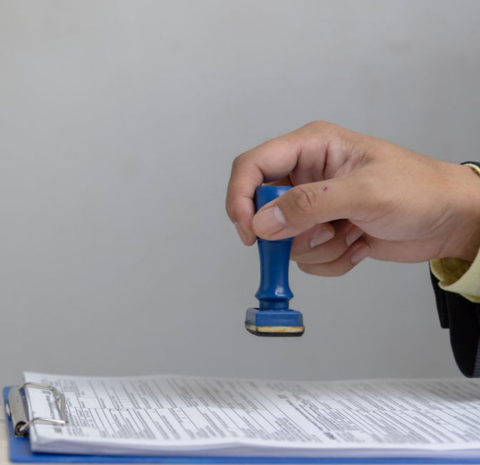 Hand holding a blue rubber stamp over a document on a clipboard. The setting is neutral, focusing on the action of stamping paperwork.