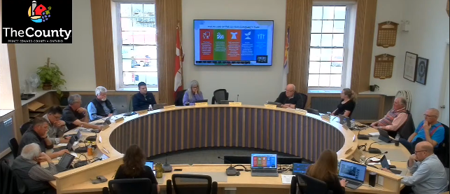 People seated around a circular table in a meeting room. A screen displays colorful charts. The room has large windows and flags.