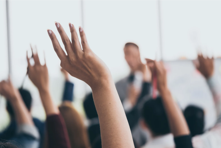 Raised hands in a bright classroom as a blurred presenter speaks, suggesting eager participation.