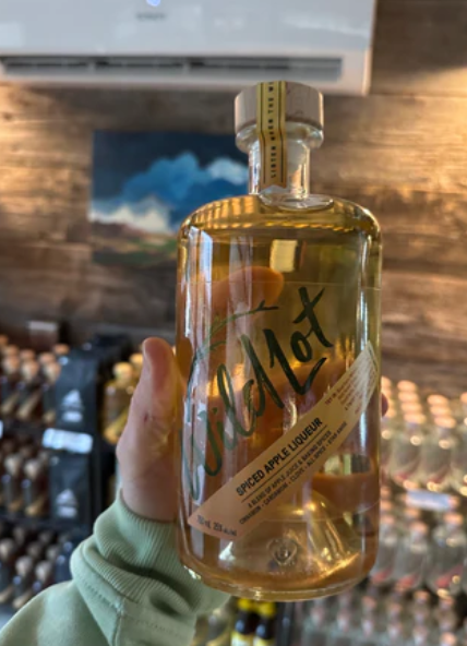Hand holding a bottle of spiced apple liqueur with "WildLot" text, set against a rustic wooden backdrop and shelves of bottles.