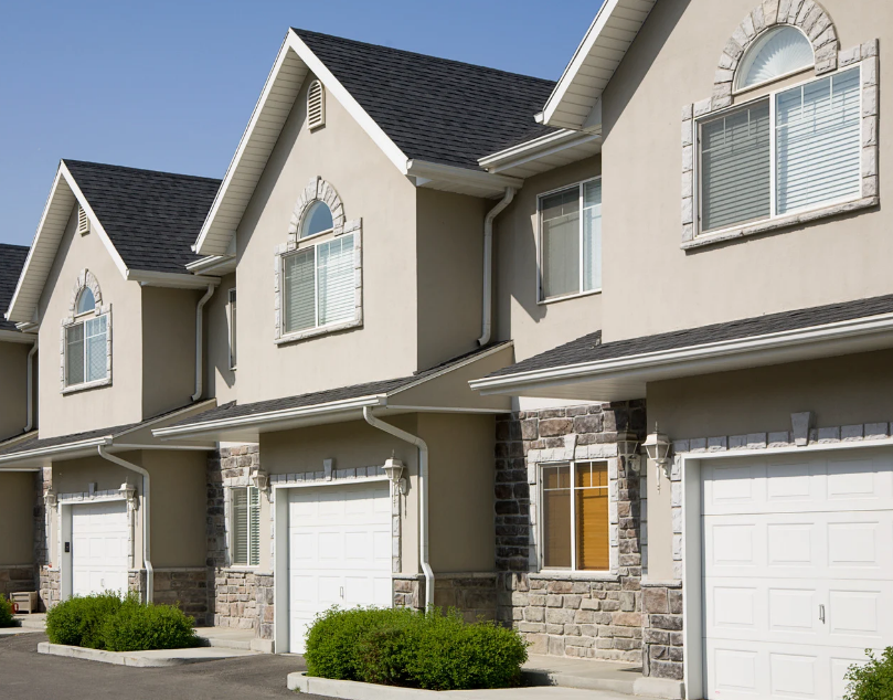 Row of beige townhouses with white garages and stone accents under a clear blue sky, with neatly trimmed shrubs.