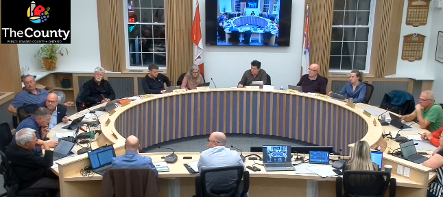 People sit around a large circular table in a meeting room. Laptops are open, and a screen displays an image. "The County" logo is visible.