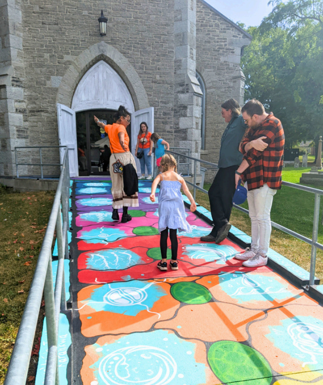 People on a colorful ramp in front of a stone building. A child walks on bright patterns; adults watch. The mood is lively and cheerful.