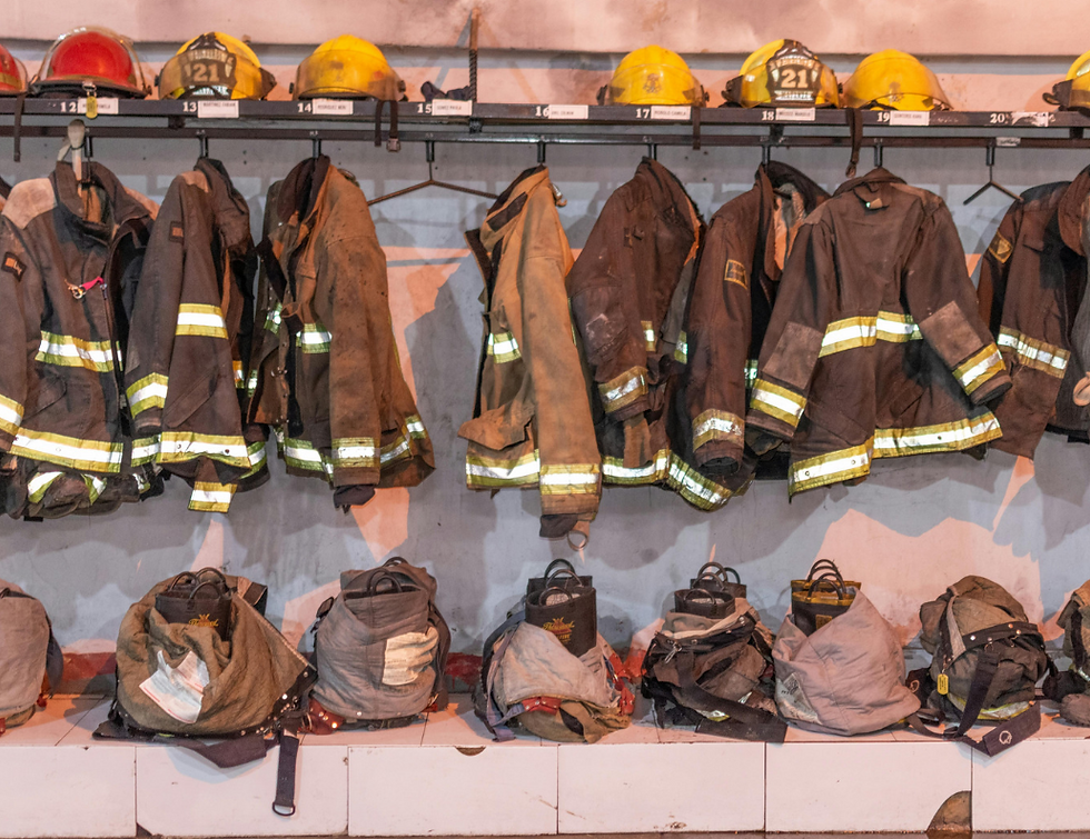 Firefighters' gear hangs neatly in a row. Jackets with reflective stripes and yellow helmets are above stacked bags on a simple white shelf.