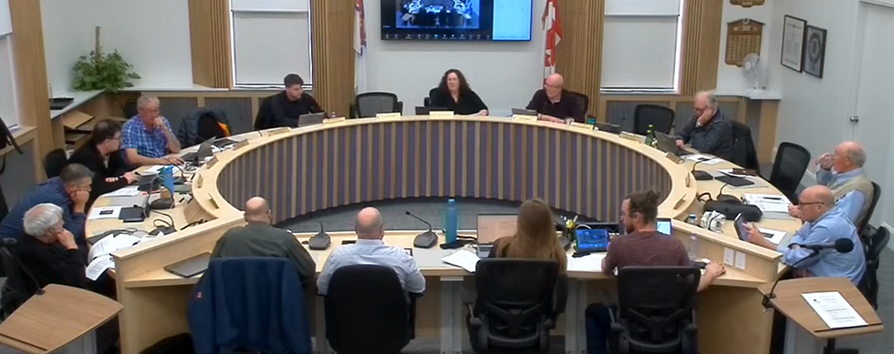 Council meeting with 12 people seated around a curved table. Laptops and papers are present. Flags and a screen in the background.