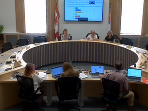 A round table meeting with nine people, some with laptops. A screen displays charts and text. Canadian flag in the background.