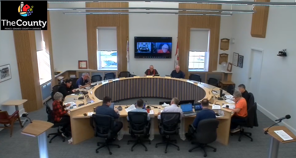 People seated around a curved table in a conference room, engaged in discussion. A screen displays video calls. Logo: "The County."