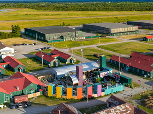 Aerial view of a colorful outdoor event with people gathered. Green buildings with red roofs surround the area amidst vast green fields.