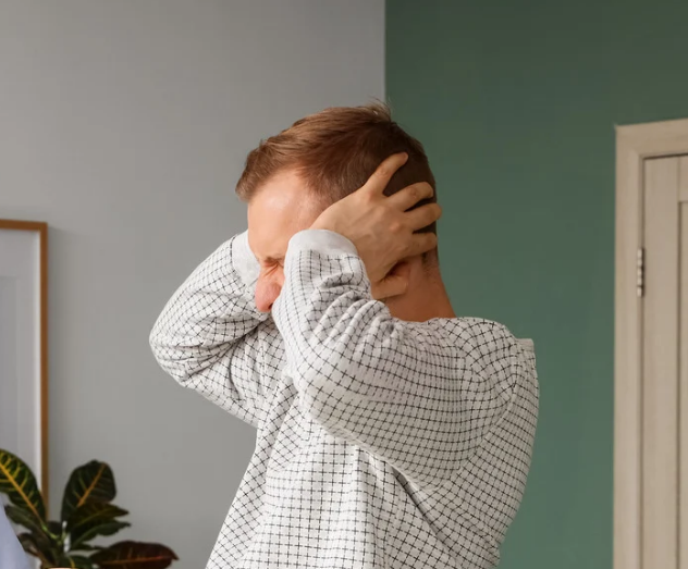 A person in a checkered shirt holds their head with both hands, appearing distressed. They stand in a room with pale green walls and a leafy plant.