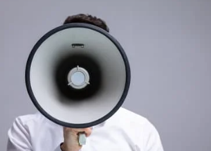 Person in white shirt holding a gray megaphone to their face against a plain background, emphasizing communication or announcement.