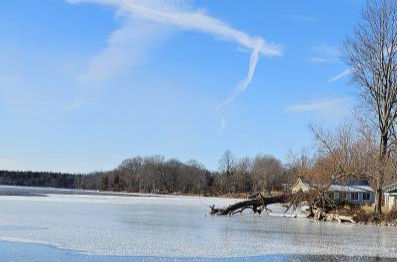A wide shot of a frozen, snow-covered lake or bay under a bright blue sky with a few contrails. A fallen tree lies near the shore next to a small white house.