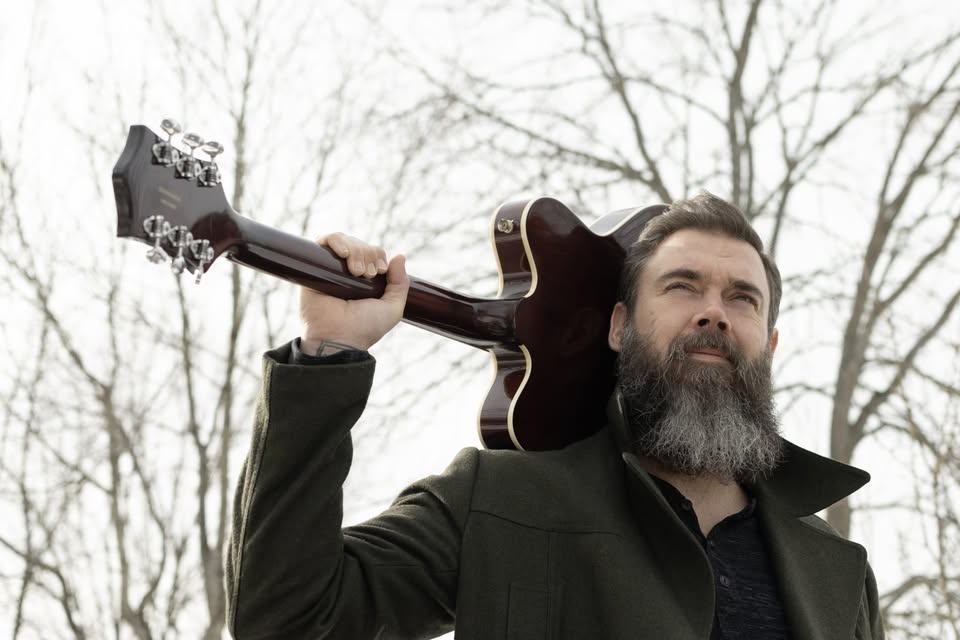 Bearded man in dark coat carries an electric guitar over his shoulder outdoors, with bare trees and a moody winter sky.