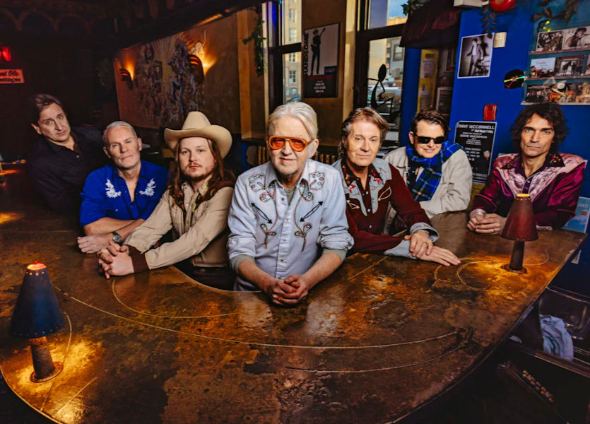 Six men in cowboy and retro shirts sit at a bar in a warm, rustic room with posters, posing calmly for the camera.