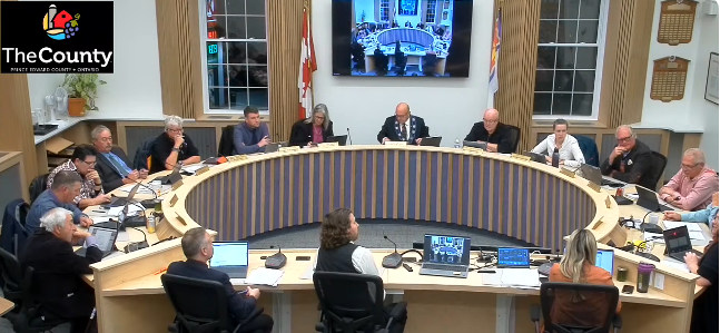 A round table meeting with 13 people. Screens, notebooks visible. Flags and a large screen in the background. Logo: "The County."