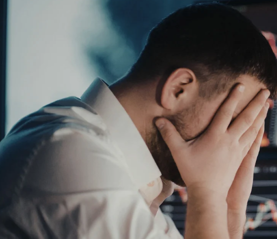 A man in a white shirt covers his face with his hands, appearing stressed. A blurred computer screen with graphs is in the background.