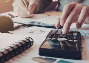 Hands using a calculator and writing in a notebook on a desk with charts. The scene suggests focus and analysis in a workspace.