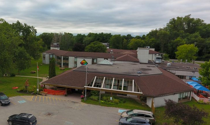 Aerial view of a building with brown roofs, surrounded by green trees. Cars in parking lot, colorful flag, and cloudy sky. Calm atmosphere.