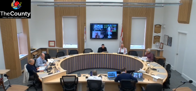 Meeting in a circular wood-paneled room with six people seated at desks, laptops open. Screen displays a video call. Logo reads TheCounty.