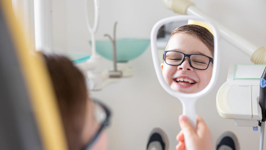 Glücklicher Junge mit Brille lacht in den Handspiegel beim Zahnarzt.