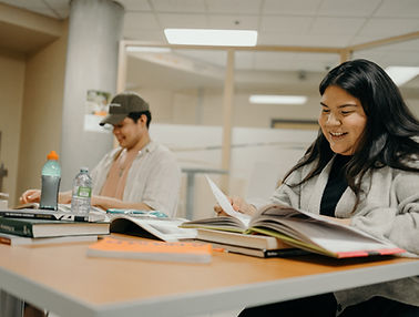 Two students seated together reading books and smiling.
