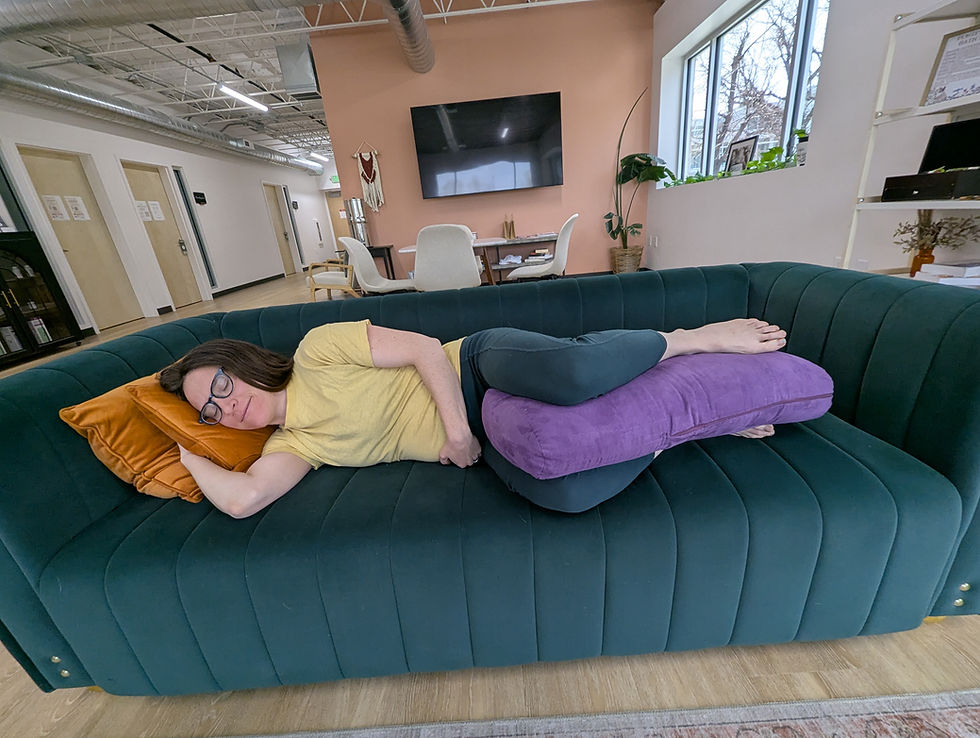 Person in a yellow shirt sleeping on a green couch with an orange pillow under their head and a purple pillow between their legs in a modern room.