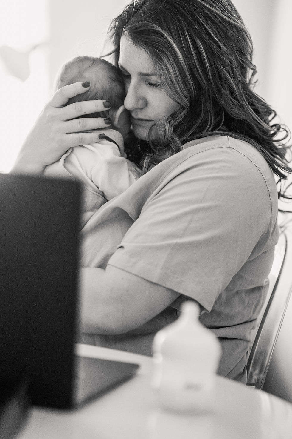 Black-and-white close-up of a woman cradling a newborn against her face, eyes closed, sitting in front of a computer and baby bottle.