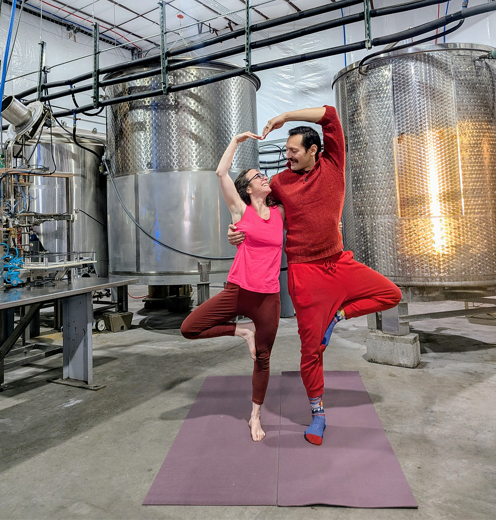 Two people in a winery, posed in a yoga tree pose on mats, smiling. They're wearing red outfits, with large metal tanks in the background.