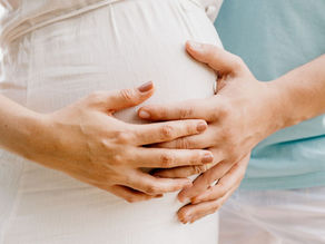 Hands gently rest on a baby bump, showing tender connection. The woman wears a white dress; background is soft blue. Maternity theme.
