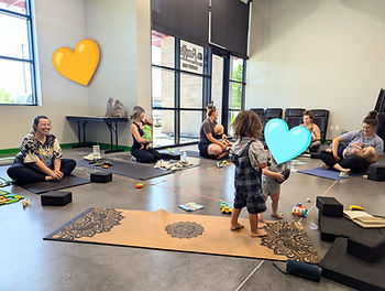 Moms with babies and toddlers in a bright studio, sitting on mats during a playful class; smiley, relaxed mood.