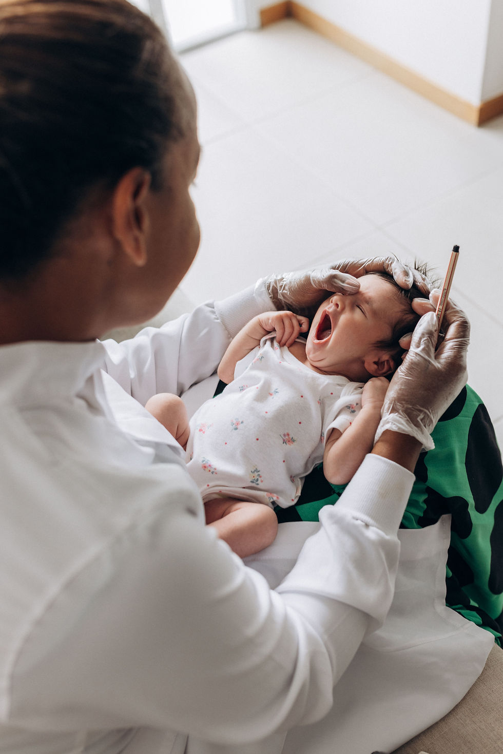Caretaker in white gloves examines a yawning baby with a thermometer in a bright clinic.