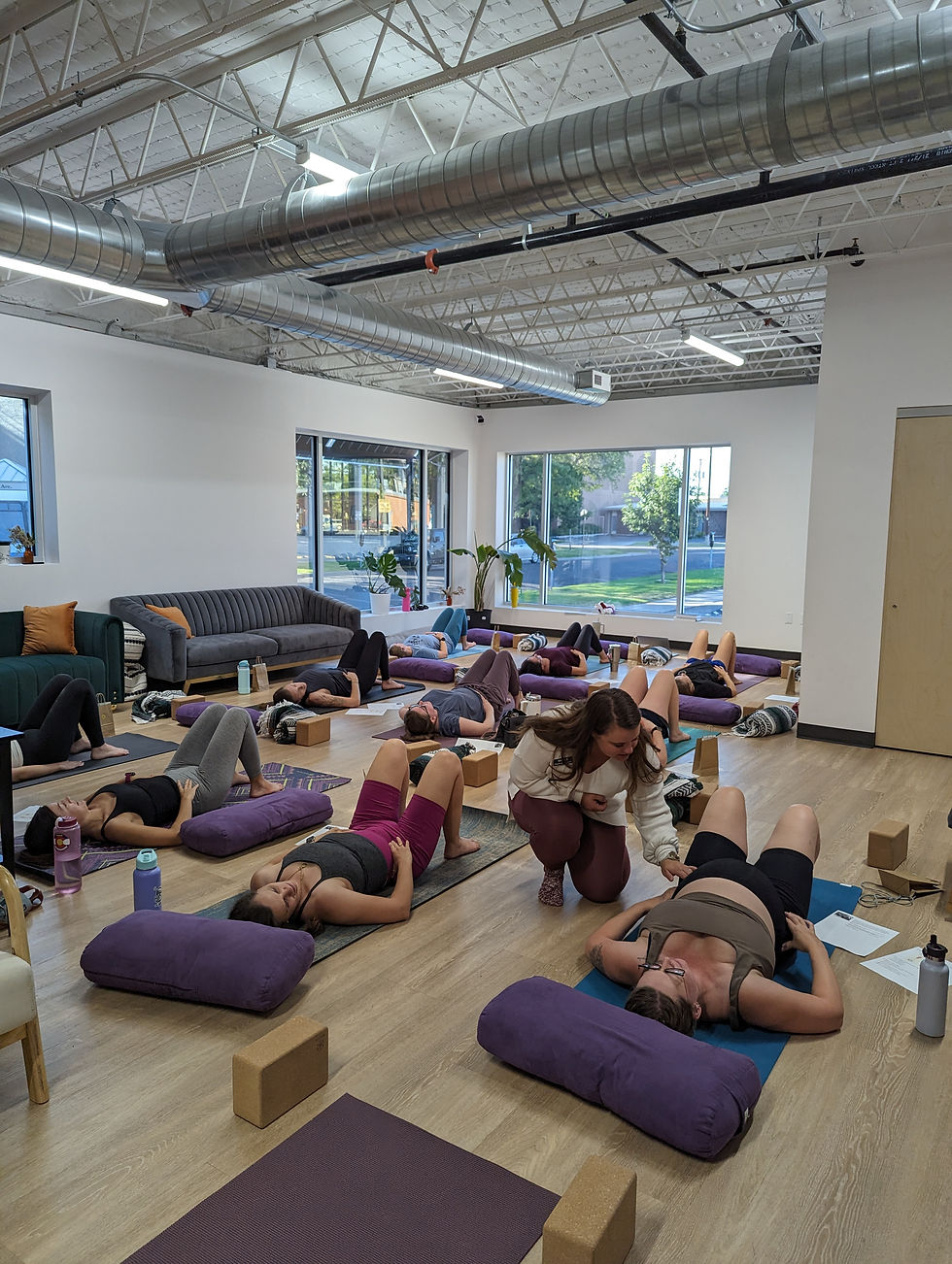 Pelvic floor physical therapist helps students in a bright studio with exposed ducts, mats, bolsters, and large windows.