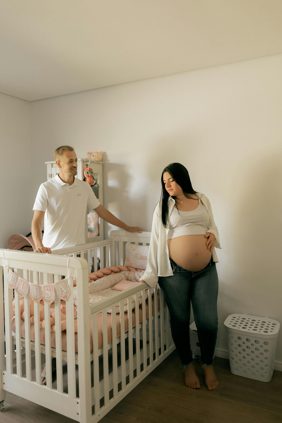 Pregnant woman stands beside a white crib labeled ELISA while her husband smiles in a softly lit nursery.