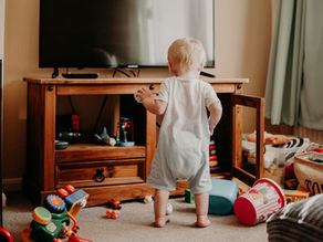 Toddler in white onesie stands by a TV cabinet in a toy-filled living room, reaching for a small ball on the wooden shelf.