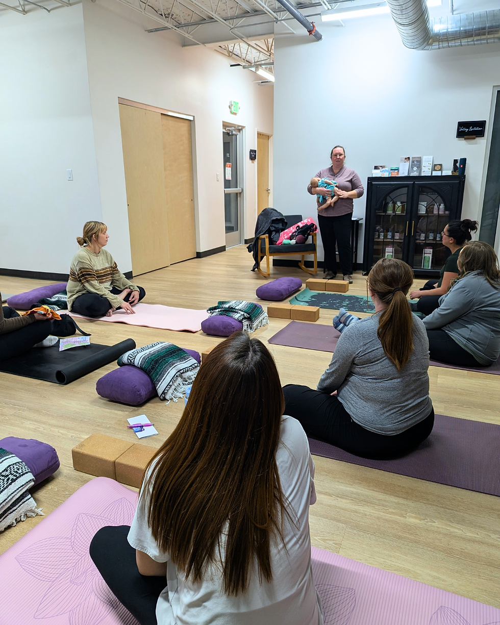Women sit on yoga mats in a bright studio while a lactation specialist holds a baby doll and speaks during a calm wellness class