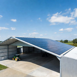 Hangar agricole moderne en France équipé de panneaux solaires photovoltaïques sur toute la toiture, tracteur stationné devant le bâtiment, champs verdoyants et ciel bleu, photographie réaliste d’une exploitation agricole durable et énergie solaire