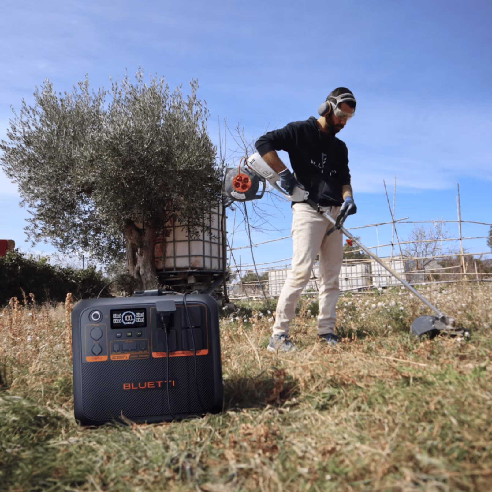 Station électrique BLUETTI Premium 200 posée au sol dans un champ, alimentant du matériel pendant qu’un utilisateur travaille au coupe-herbe.