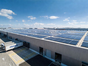 Photographie ultra réaliste d’un grand entrepôt logistique moderne avec une immense toiture couverte de panneaux solaires photovoltaïques, zone industrielle française avec camions et quais de chargement, panneaux solaires brillants au soleil, ciel bleu lumineux, style photographie d’architecture industrielle durable, énergie renouvelable pour entreprises, rendu 8k très détaillé, profondeur de champ réaliste