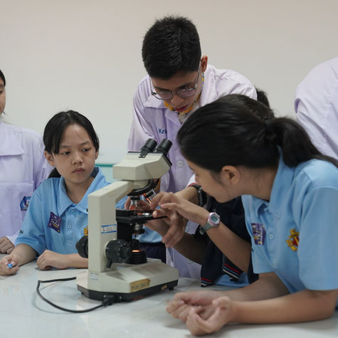 SDH Lippo Village Students in lab coats and blue uniforms study a microscope together in a classroom lab, focused and curious.