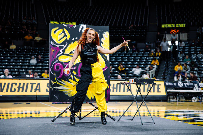 Speed painter Annika Wooton performing live during a Wichita State women’s basketball halftime show in Wichita, Kansas, painting on a black canvas on the arena court.