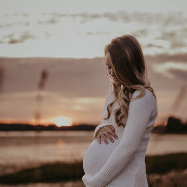 Photo of a woman in a white dress holding her pregnant belly at sunset on a beach.