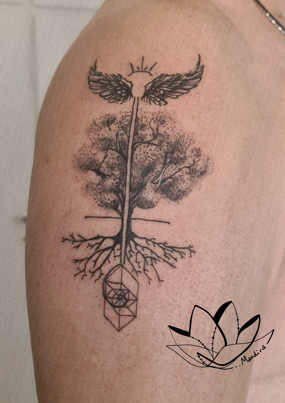 black and white tattoo on a male upper arm / shoulder, tree of life and crystal spiral, angel wings and the sun rays on the top. Mandira Tattoo & Art logo on the right bottom.