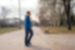 A blind man with a guide cane walks along a paved path in a park, dressed in a blue jacket and jeans. the autumn leaves and trees create a peaceful outdoor atmosphere.
