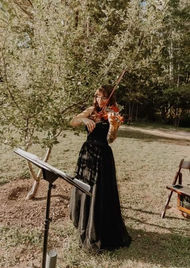 First String Violinist stands in the shade of a small tree in Utah, dressed in a floor length black dress, playing the violin for a bride