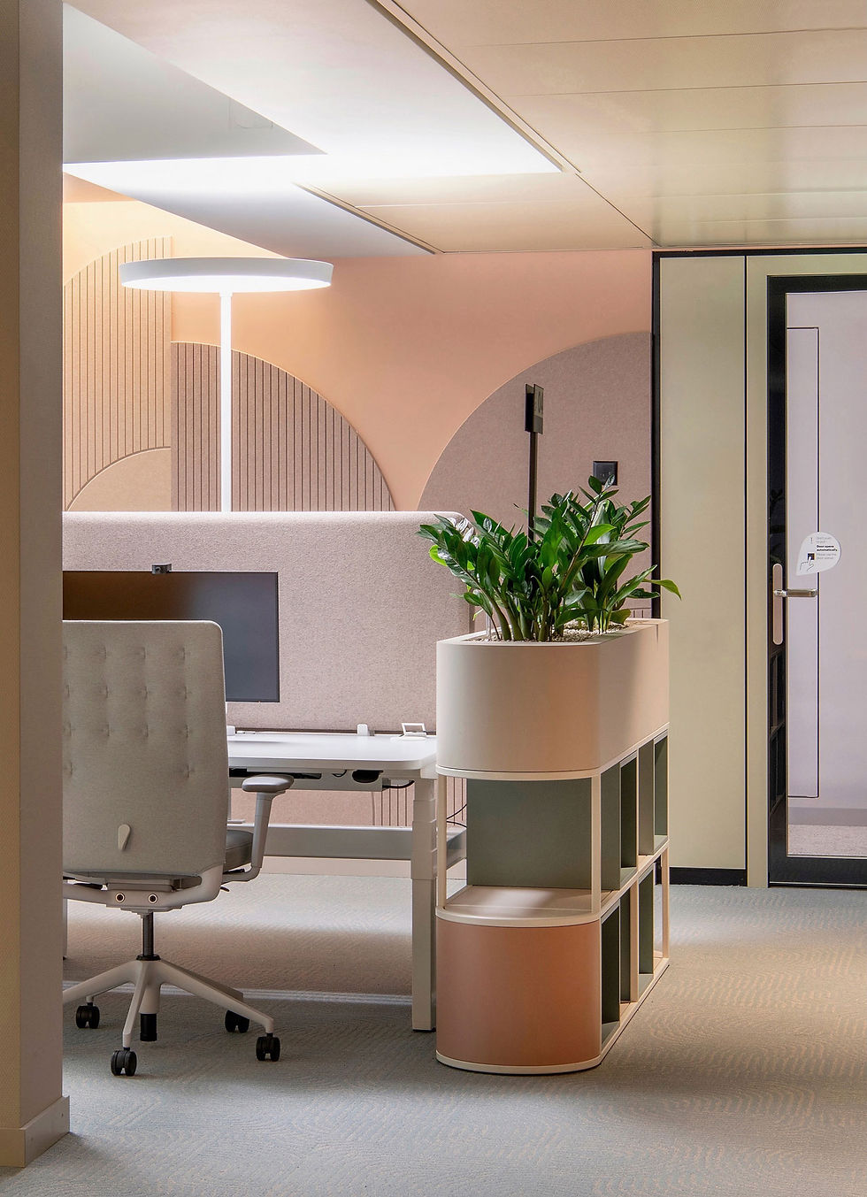 A modern office workspace featuring a ZZ Plant (Zamioculcas zamiifolia) in a sleek planter beside a desk. The setting includes soft lighting, pastel tones, and contemporary furniture creating a calm, natural atmosphere.