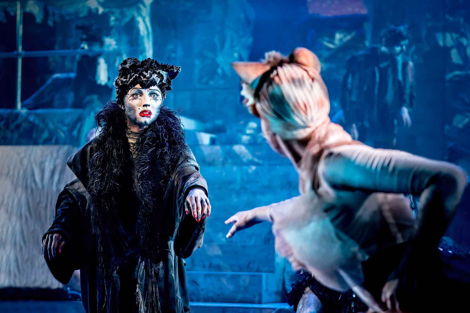The image captures a dramatic theatrical scene featuring two performers in CATS in elaborate, stylized costumes. One performer, dressed in dark attire and heavy makeup, appears to be the central focus, while the other, with cat-like features, gestures toward them.
