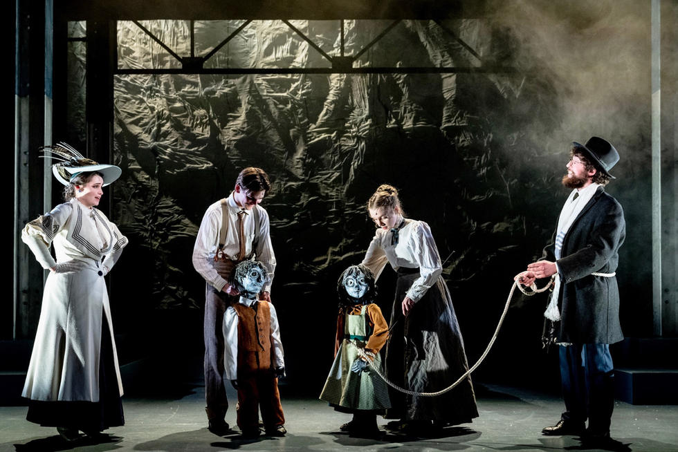 Image shows actors at Arts Ed, during a performance of Ragtime the musical. Four human actors playing adults, with two puppets playing children stand in a line on stage in period costumes. One of the puppets is tethered to an actor by a long piece of rope.