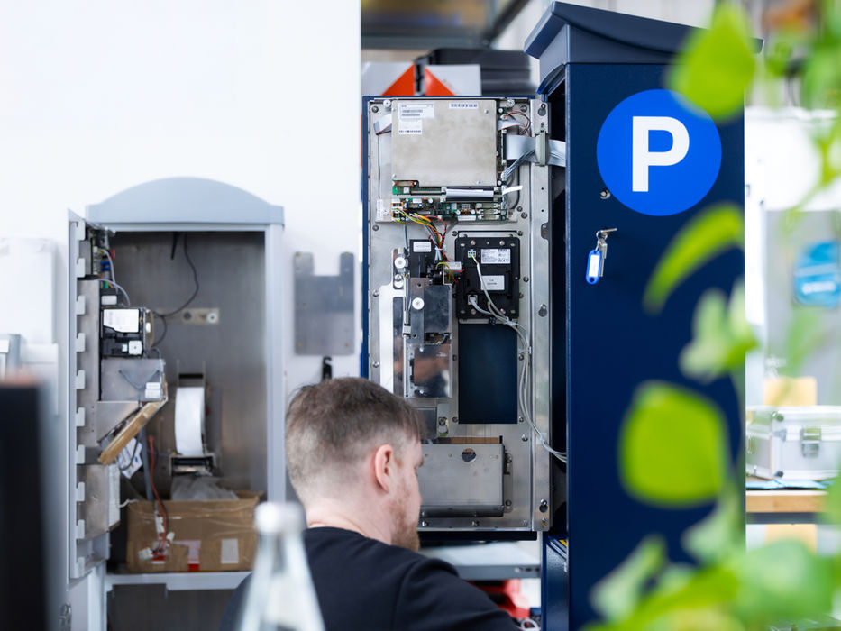 Mann repariert Parkautomat mit "P"-Symbol, interne Komponenten sichtbar.