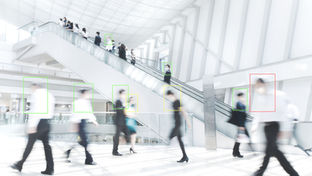 Blurred crowd of people walking through a bright modern building with an escalator, highlighted by tracking boxes illustrating retail visitor analytics and people counting technology.