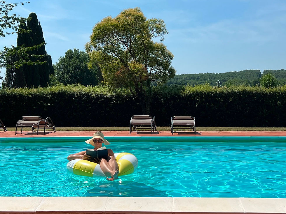Woman in sunhat reads on a yellow-and-white float in a bright blue pool, with lounge chairs and trees behind.